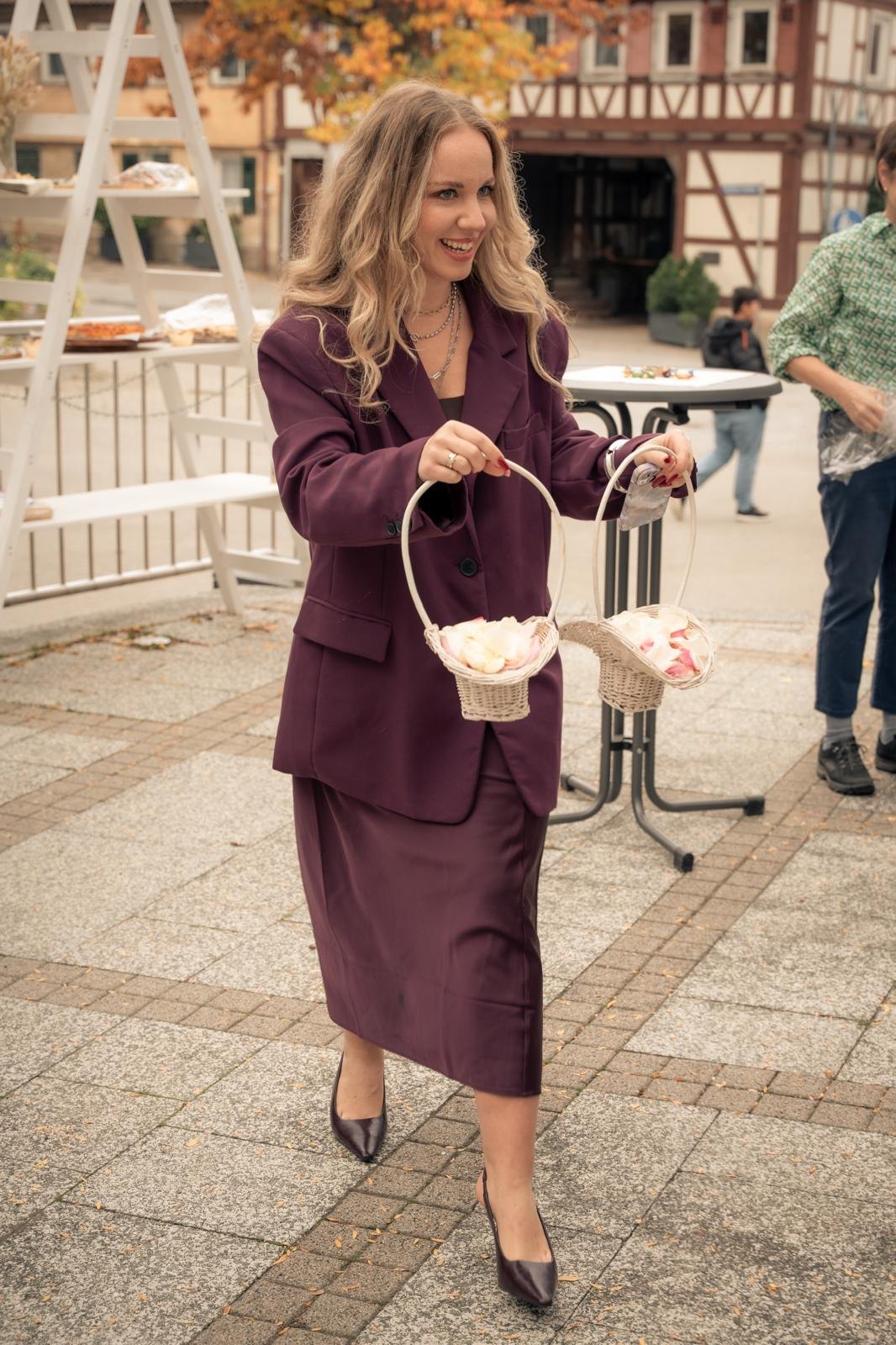 Sabrina trägt lachend zwei Körbe mit Blütenblättern über einen Hof